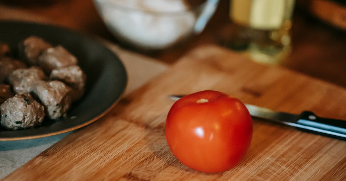 High angle of fresh ripe tomato and knife on wooden chopping board near plate with meat and oil in glass bottle near bowl on counter in kitchen